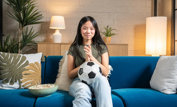 Asian woman watching football, holding a soccer ball, cheering while sitting on a couch