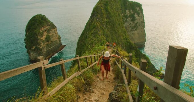Woman climbing up narrow steep rocky path with wooden rails with ocean view at Kelingking Beach Bali Nusa Penida