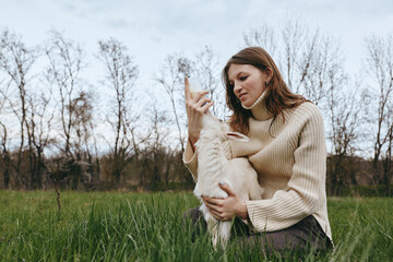 Naklejka premium Woman in a cozy sweater sits in a green meadow, embracing a fluffy goat kid, sharing gentle affection outdoors, conveying wholesome lifestyle moments and natural bond with animals.
