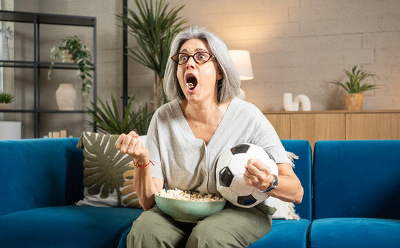 Enthusiastic senior woman cheering, holding soccer ball and eating popcorn at home