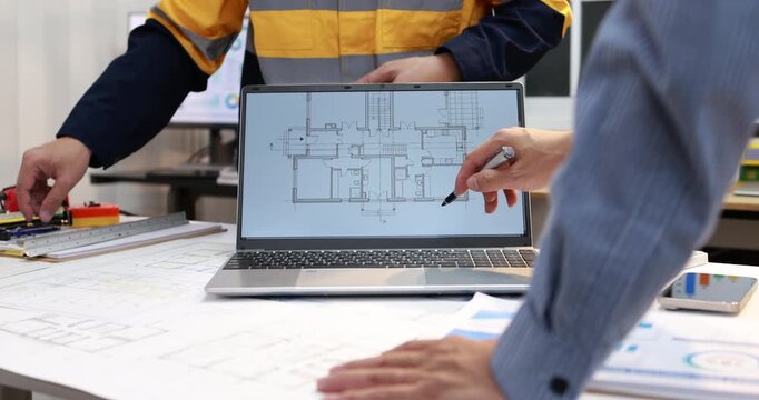 Architects review a building floor plan on a laptop while discussing printed drawings at an office desk.