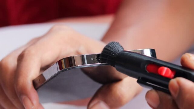 Asian man's hand cleaning a smartphone speaker grille with a small cleaning brush, also brushing around the charging port to remove dust, lint, and debris for better device maintenance and care.
