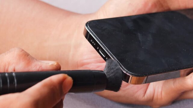 Asian man's hand cleaning a smartphone speaker grille with a small cleaning brush, also brushing around the charging port to remove dust, lint, and debris for better device maintenance and care.
