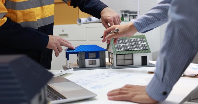 Architects and construction workers discuss two modern house models over building plans in a bright office, planning a residential project.