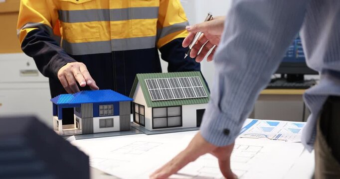 Architects and solar technician discuss home plans with model houses, including a rooftop solar panel, in a modern office.