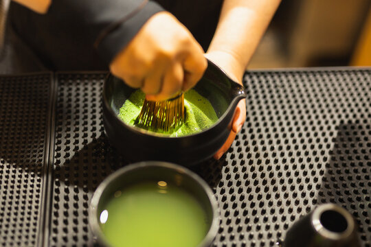 Barista whisking matcha green tea using a traditional bamboo whisk in ceramic bowl.