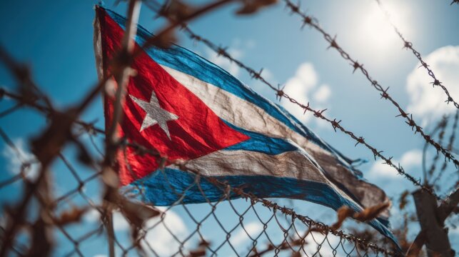 Cuban flag waving in the wind near a barbed wire fence with a clear blue sky and bright sunlight above