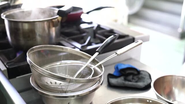 The chef is using a pot of boiling water on the gas stove to steam the spaghetti and then using a strainer to scoop it out, preparing to stir-fry the spaghetti in the kitchen.