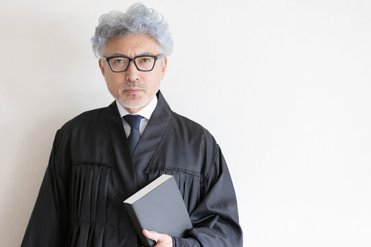 法律書を手にカメラを見つめる法服姿の男性裁判官
