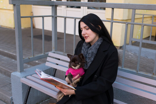 Female reader enjoying a book with her little dog on a bench against modern apartment buildings, urban outdoor lifestyle, black winter coat, autumn city atmosphere, peaceful recreation and companionsh