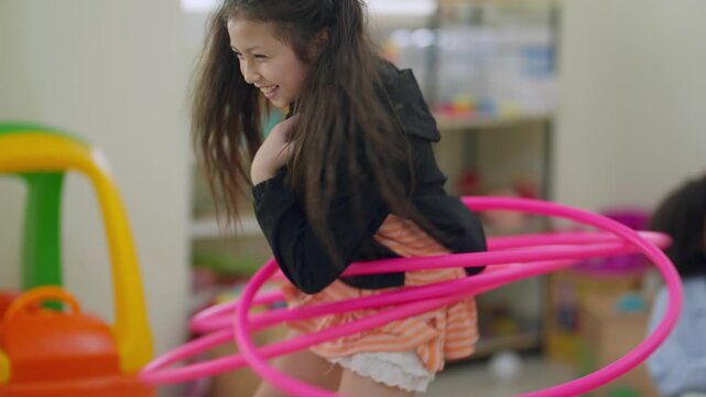 Girl laughs while spinning a pink hula hoop. Child wears striped dress and black jacket. Playroom holds colorful toys and soft light