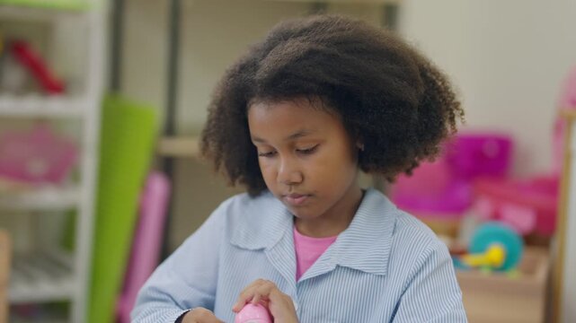 Young girl examines toy with focused interest. Curly hair frames her thoughtful expression. Playroom shelves hold colorful toys and equipment