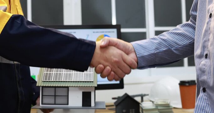 Two businessmen shaking hands over a model house in an office, a successful real estate deal, property sale, partnership, or mortgage agreement.