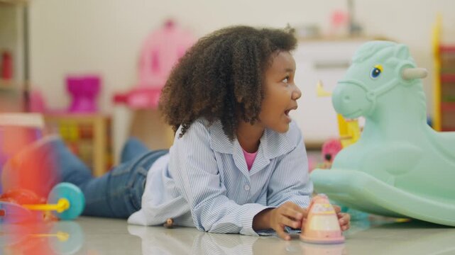 Young girl examines toy with focused interest. Curly hair frames her thoughtful expression. Playroom shelves hold colorful toys and equipment