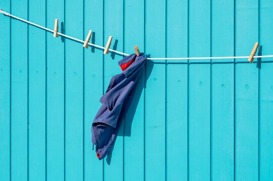 Dunkelblaue Kleidung h&auml;ngt an einer W&auml;scheleine vor einer leuchtend t&uuml;rkisfarbenen Holzwand im intensiven Sonnenlicht und erzeugt starke Schatten.