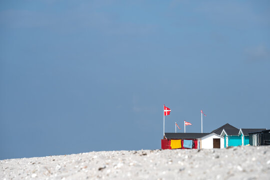 Bunte Strandh&auml;user mit wehenden d&auml;nischen Flaggen stehen &uuml;ber einem hellen Sandstrand in Marstal unter weitem blauem Himmel an einer sonnigen K&uuml;ste.