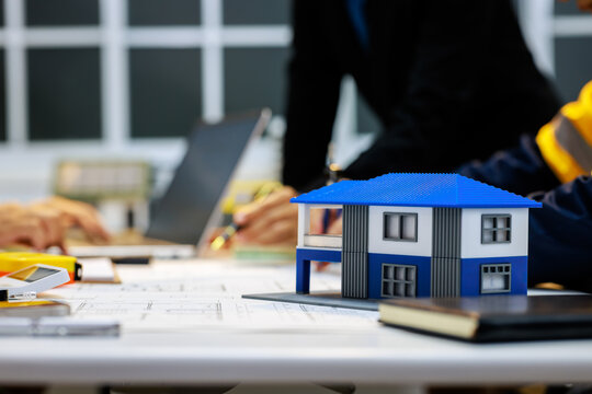 Small blue and white house model on architectural plans with engineers discussing a building project in a modern office.