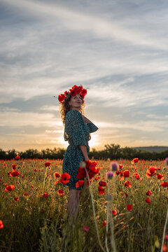 Golden hour portrait of young woman wearing Papaver rhoeas floral wreath standing in red poppy field during summer sunset