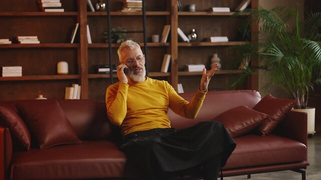 Annoyed man in his 50s argues during phone call while sitting on sofa at home