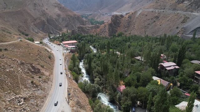Curving Chalus Road descends a brown mountain slope toward a green river valley with houses, trees, and a moving vehicle. Ideal for travel, tourism, and nature stock Iran Alborz mountains.