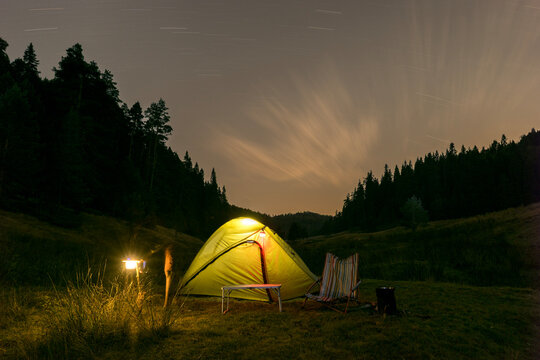 A bright yellow glowing tent set up at a night campsite surrounded by dark pine forest. A campsite scene featuring a camping light, folding chair, table, and long exposure star trails across the cloud