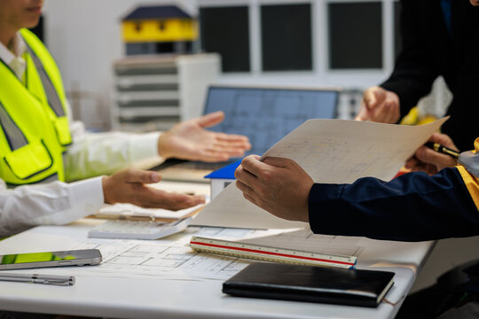 Construction professionals in safety vests review building plans at a desk with a tablet, notebook, and scale ruler in a modern office.
