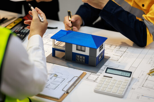 Engineers and builders discuss house plans around a model home, calculator, and blueprints during a construction planning meeting.