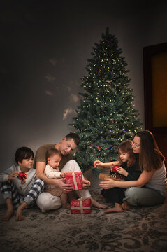 A large family of three children, mom and dad, are celebrating New Year's and Christmas under the Christmas tree at home, opening presents. A quiet, cozy family evening. vertical