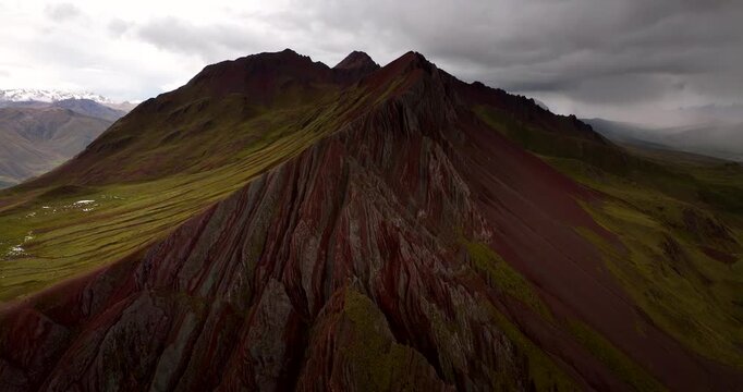 Rugged Pallay Punchu Rainbow Mountain ridges under dramatic clouds in Peru, drone aerial approach with sunlit slopes and eroded draping cliff
