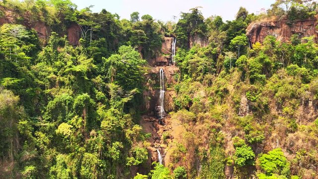 Drone view looks up at three cascading levels of Manchewe Falls, then lowers to reveal the dramatic forested cliffs near Livingstonia, Malawi.