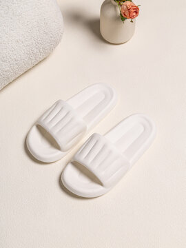 Minimalist white rubber slides or slippers on a textured surface with a towel and flower