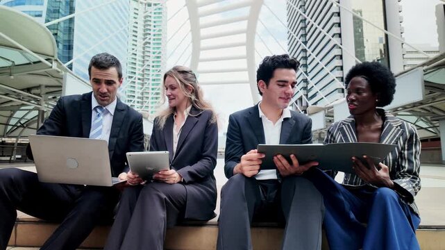 International business professionals from diverse ethnic backgrounds sitting together on city steps, discussing ideas and using digital devices in an urban setting