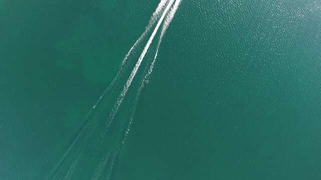 Top down drone view captures parallel white boat wakes sweeping across clear emerald water and intersecting faint trails below