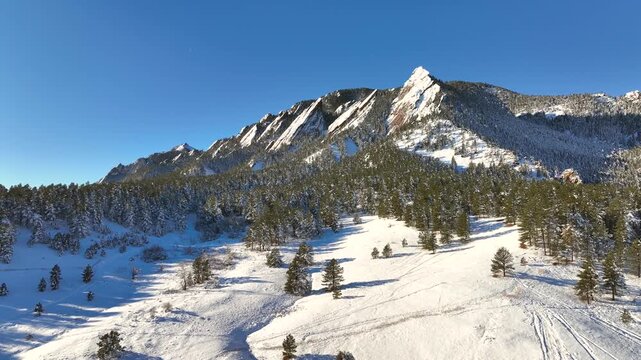 Aerial drone flight over snowy mountain landscape with rocky peaks and pine forest under clear blue sky.