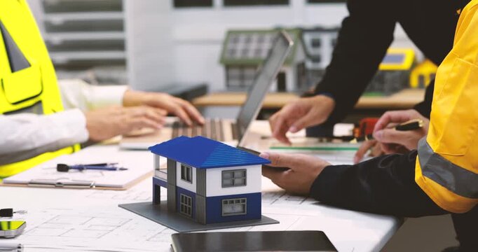 Construction team reviews plans around a small house model, laptop and tools in a modern office, showing home design, building planning and teamwork.