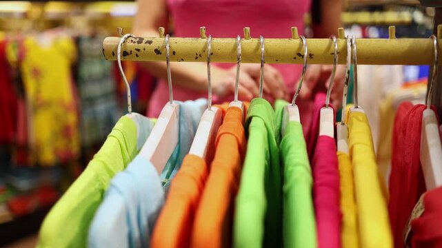 Close-up of a woman hands browsing, touching and choosing colorful clothes, tops and shirts on hangers from racks in a clothing store or boutique. Retail shopping, fashion and apparel concept.