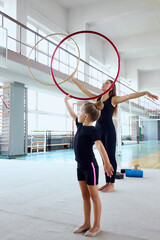 Female coach guides movement of young girl performing rhythmic hoop pose. Concept of graceful body coordination, supportive sports mentorship, and creative apparatus training. © master1305