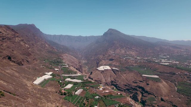 Aerial hover above a deep barranco showing terraced banana greenhouses scattered across the valley floor.