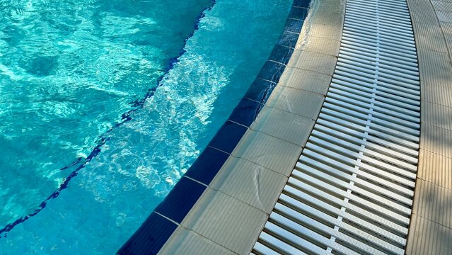 Closeup of a swimming pool edge with turquoise water, dark blue tiles and a curved white overflow drainage grate