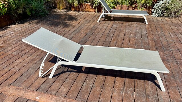 Outdoor poolside lounge chairs on a wooden deck, featuring modern white reclining sun loungers surrounded by greenery