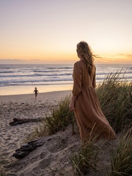Mother standing on sand dunes watching her child run on the beach during a beautiful sunset