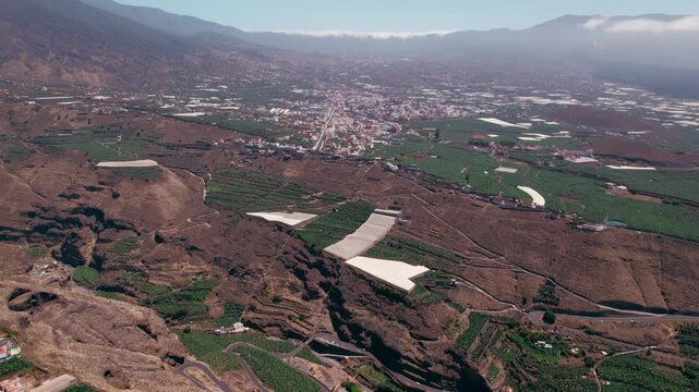 Wide drone view captures a valley town framed by terraced farmland and cloud covered mountains.