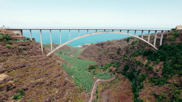 A concrete arch bridge stretches across a steep volcanic valley lined with banana terraces near the coast.
