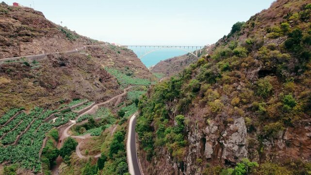 A scenic mountain valley opens toward a distant arch bridge and the shimmering Atlantic coastline.