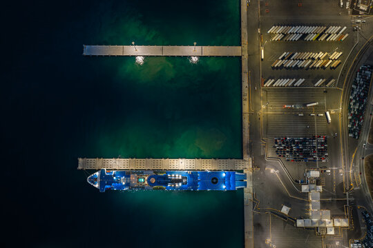 Aerial view of a large ferry docked at the pier of Porto Civitavecchia at night, illuminated by artificial lights, with rows of parked vehicles in the port area, Civitavecchia, Lazio, Italy.