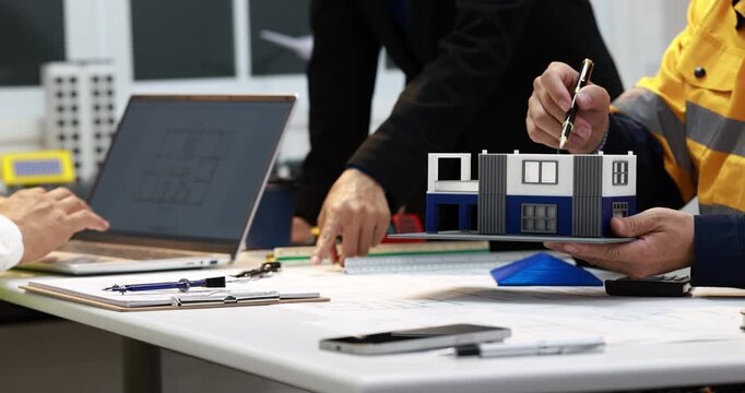 Engineers and a construction worker review plans and a small building model at a desk, teamwork, site planning and architectural design.