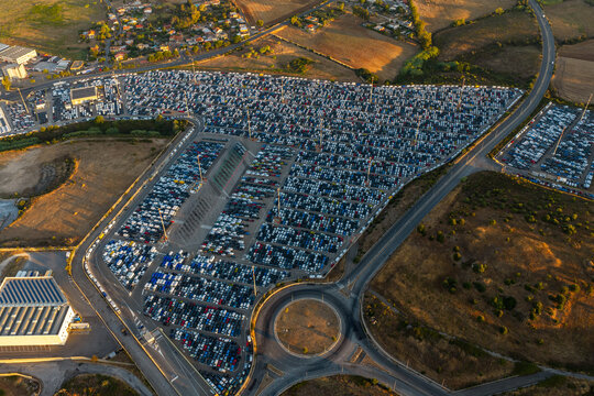 Aerial view of a large automotive storage lot filled with rows of new cars, located near a roundabout and industrial buildings in Civitavecchia, Lazio, Italy.