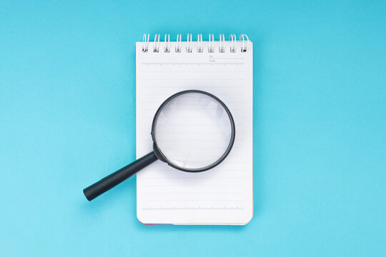 Flat Lay of Magnifying Glass Above Notebook Against Blue Background