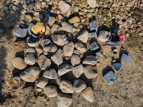 Aerial view of two workers shaping large river stones with hand tools on a sandy ground in Karanglewas, Central Java, Indonesia.