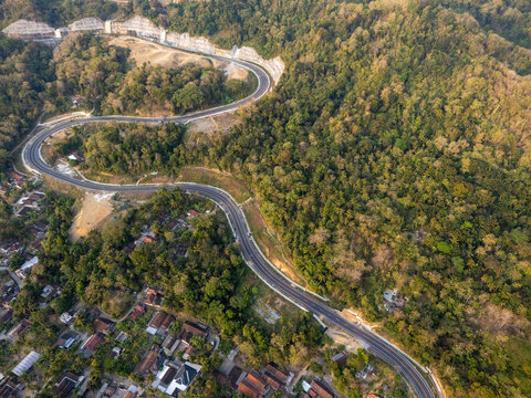 Aerial view of a winding road cutting through a lush forested landscape with scattered houses in the valley, Bantul, Special Region of Yogyakarta, Indonesia.